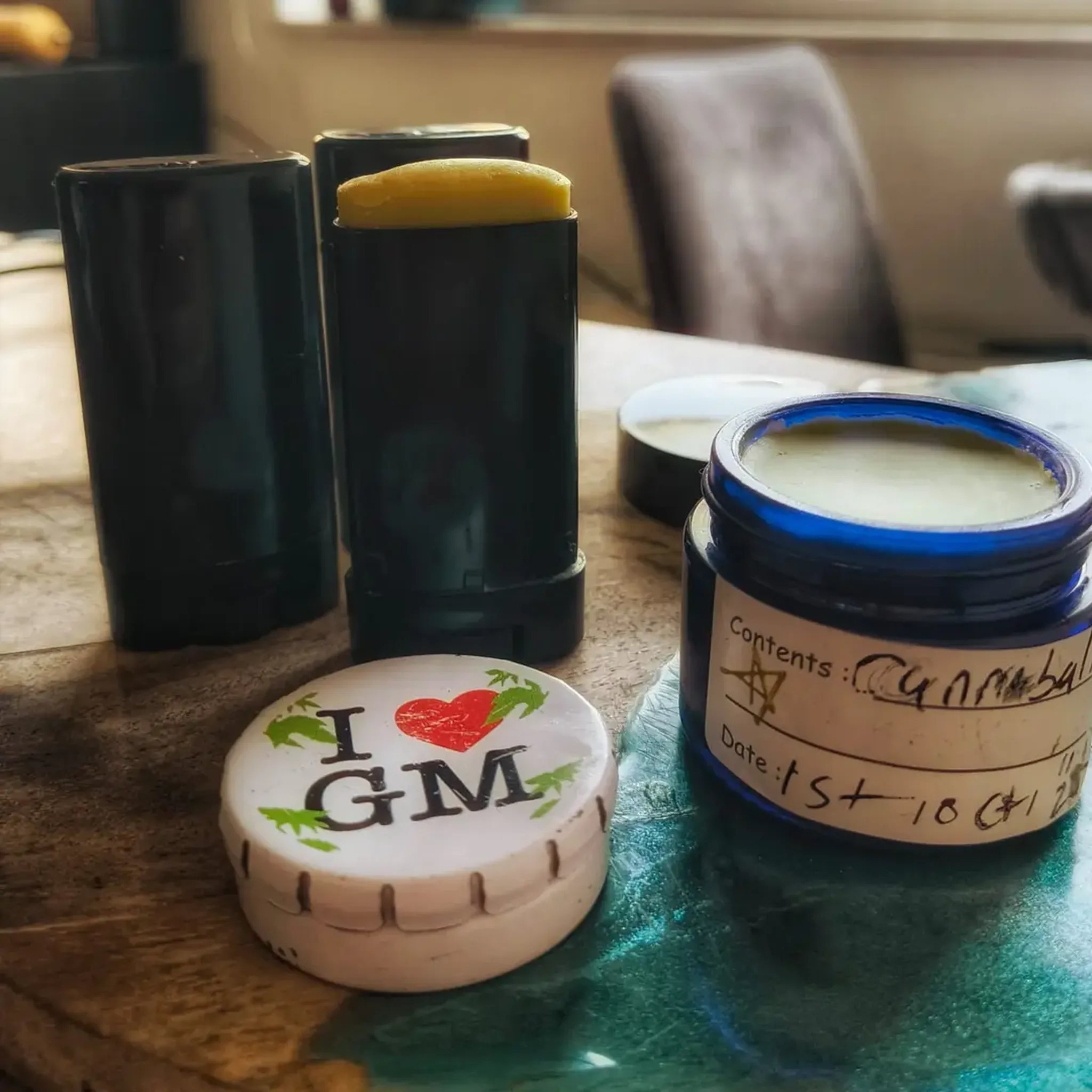 Black balm stick with yellow product, a white lid labeled "I ♥ GM", and a blue jar with a handwritten label on a wooden table.