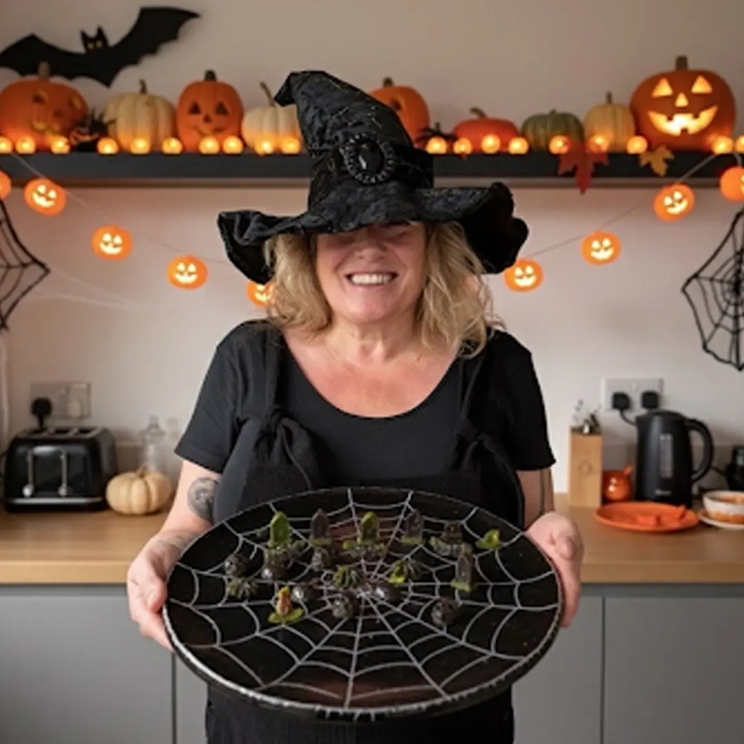 Woman in witch hat holding a platter with spider web design. Halloween decor with pumpkins and lights in the background.