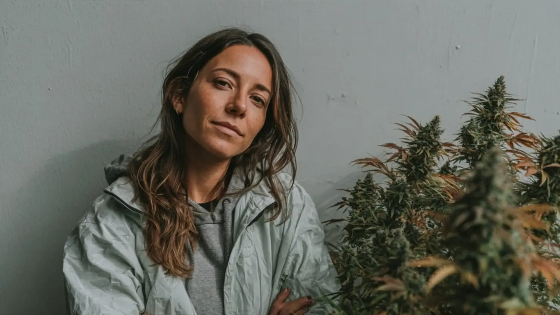Woman with long hair in a light jacket stands beside tall plants against a gray wall, looking at the camera.