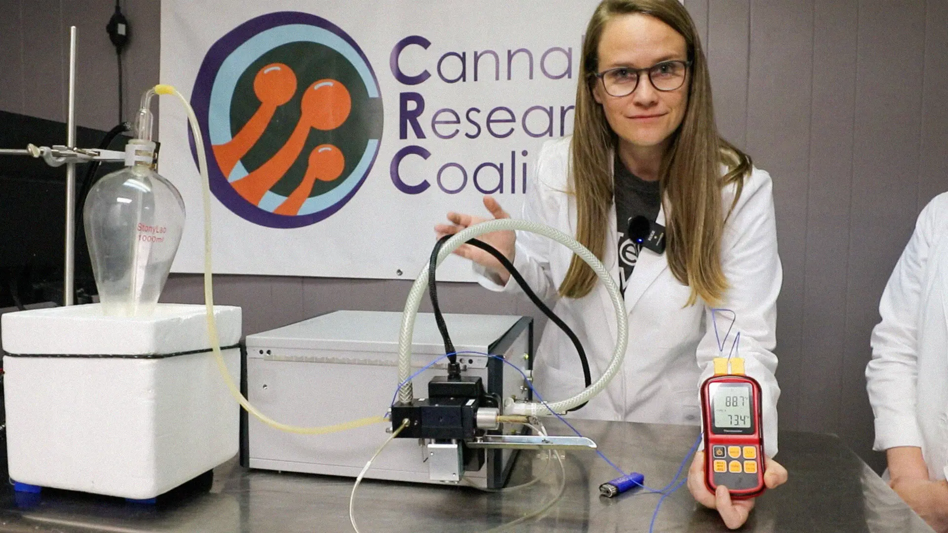 A scientist in a lab coat demonstrates equipment, holding a digital device, with a