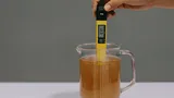 Hand holding a digital pH meter in a glass pitcher filled with brown liquid, displaying a pH reading of 6.13.