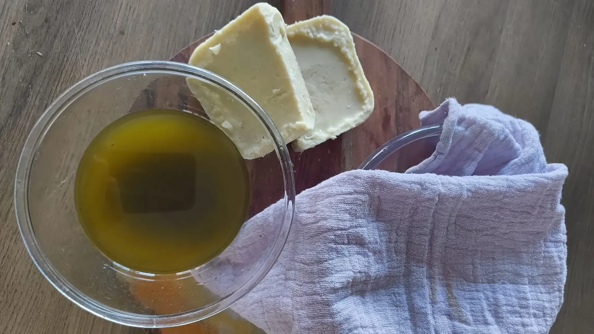 A glass bowl of olive oil, two slices of cheese on a wooden board, and a folded cloth on a wooden surface.