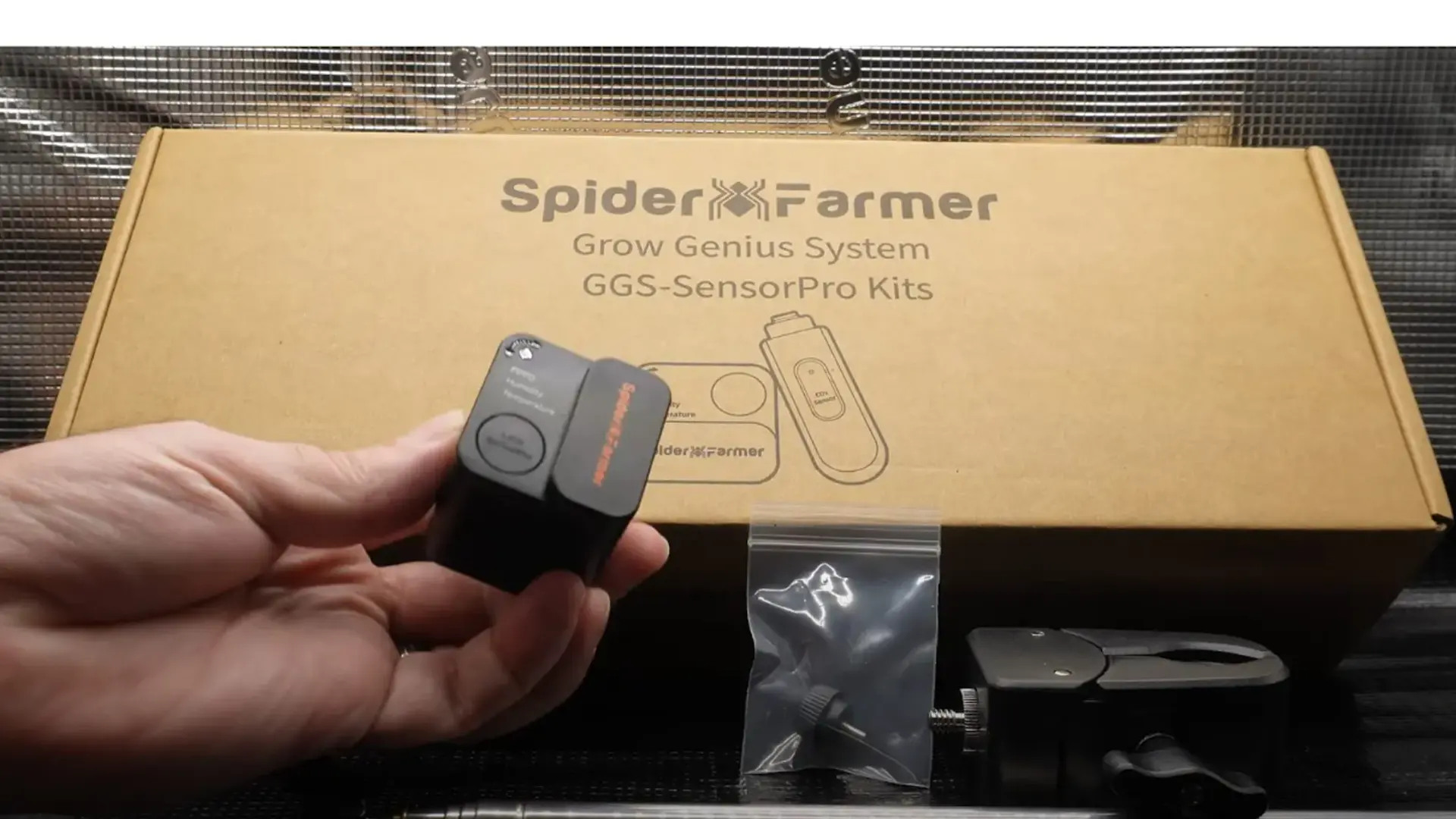 Hand holding a small device in front of a Spider Farmer Grow Genius System box, with additional components on a table.