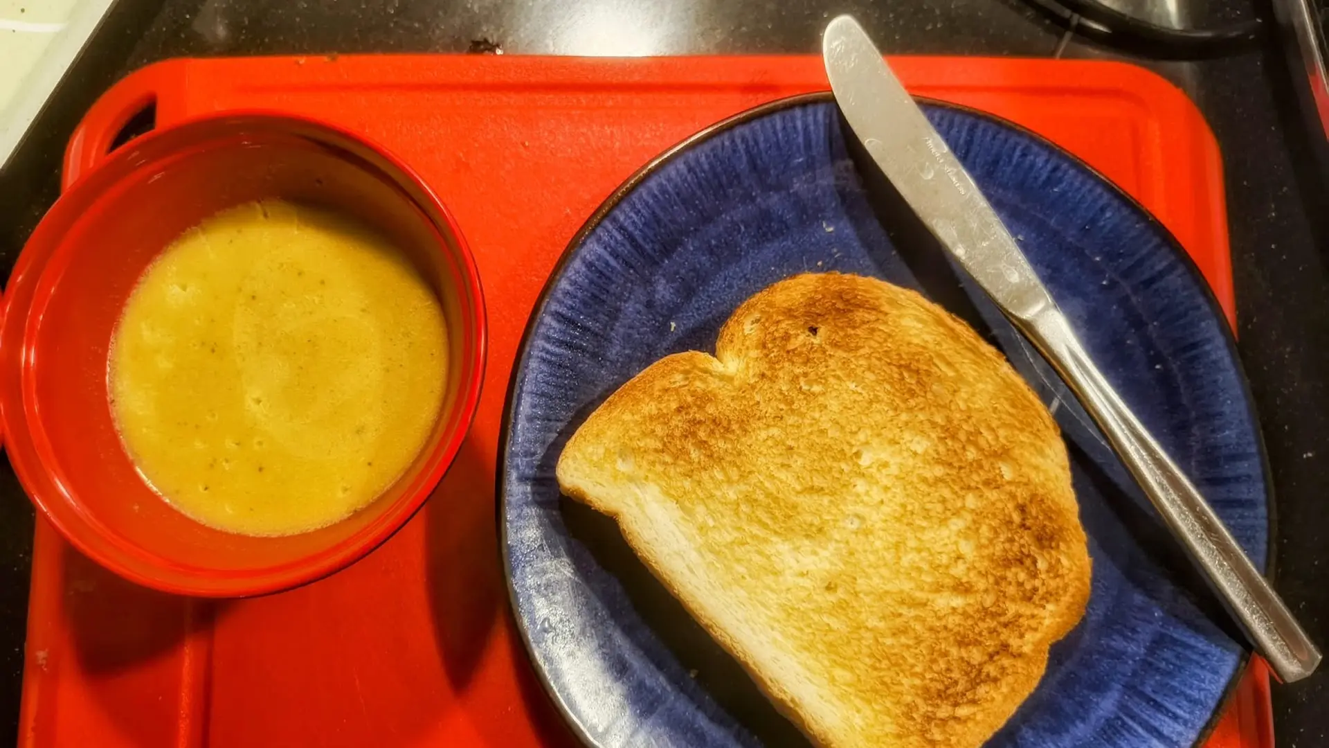 A bowl of soup and a slice of toast on a blue plate with a knife, placed on a red surface.