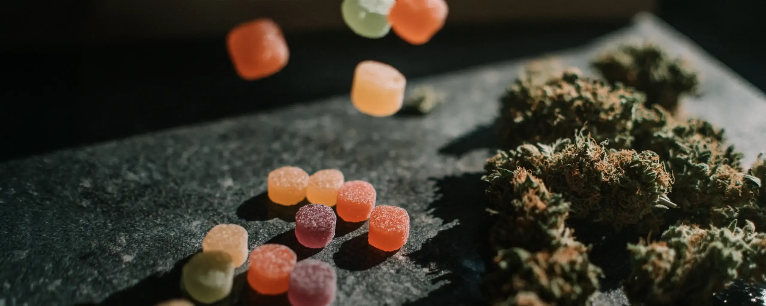 Colorful gummy candies are scattered on a dark surface next to dried cannabis buds.