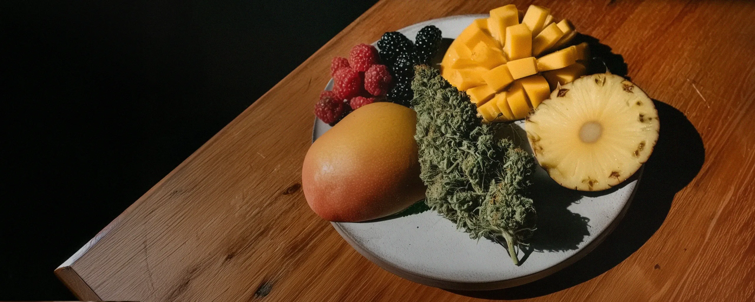 A plate on a wooden table with mango, raspberries, blackberries, sliced pineapple, and a large cannabis bud.