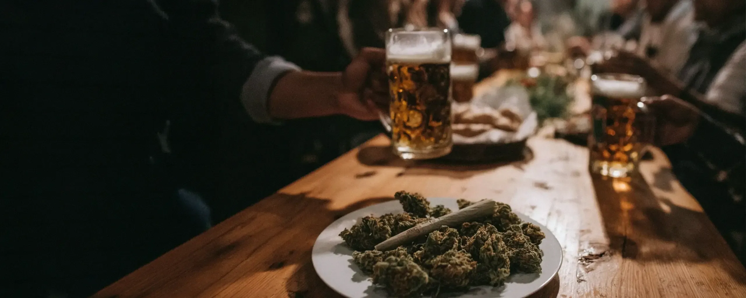 A plate with cannabis buds and a joint on a wooden table, with a hand holding a glass of beer in a dimly lit setting.