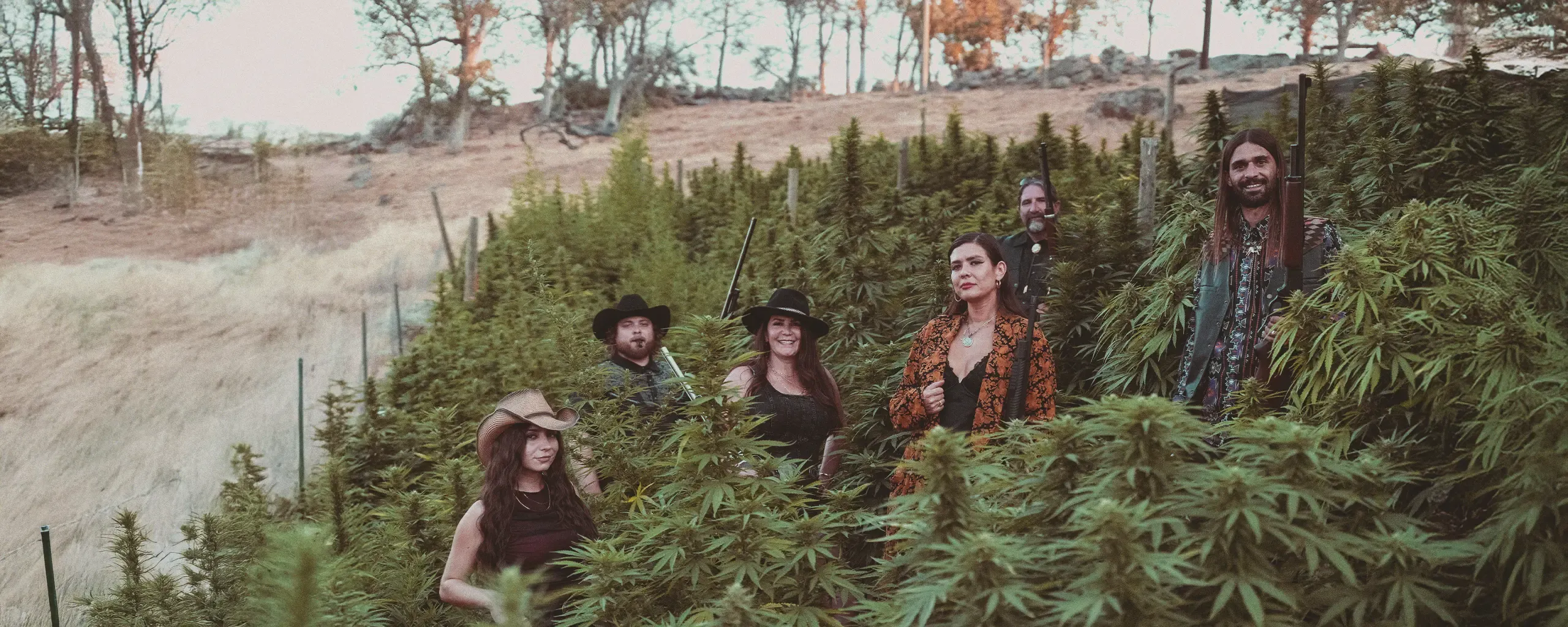 A group of people in hats and casual clothing stand among tall cannabis plants in a rural outdoor setting with trees in the background.