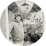 Person smiling in a greenhouse, surrounded by hanging plants and potted greenery, wearing a cap and hoodie.
