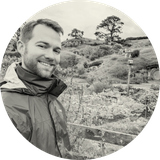 Smiling man in a rain jacket stands in a lush garden with rolling hills and trees in the background.