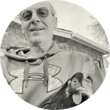 Man in glasses and hoodie smiling, holding a chicken. Brick building and tree in the background. Black and white image.