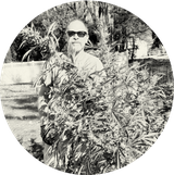 Person wearing sunglasses standing outdoors, partially obscured by tall plants, with trees and a fence in the background. Black and white image.