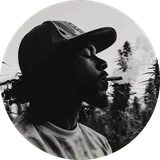 Silhouette of a person with curly hair and a cap, exhaling smoke from a cigar, surrounded by tall plants.