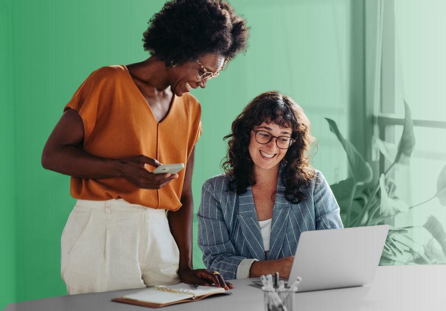 Two women smiling while reviewing a laptop and phone at a desk