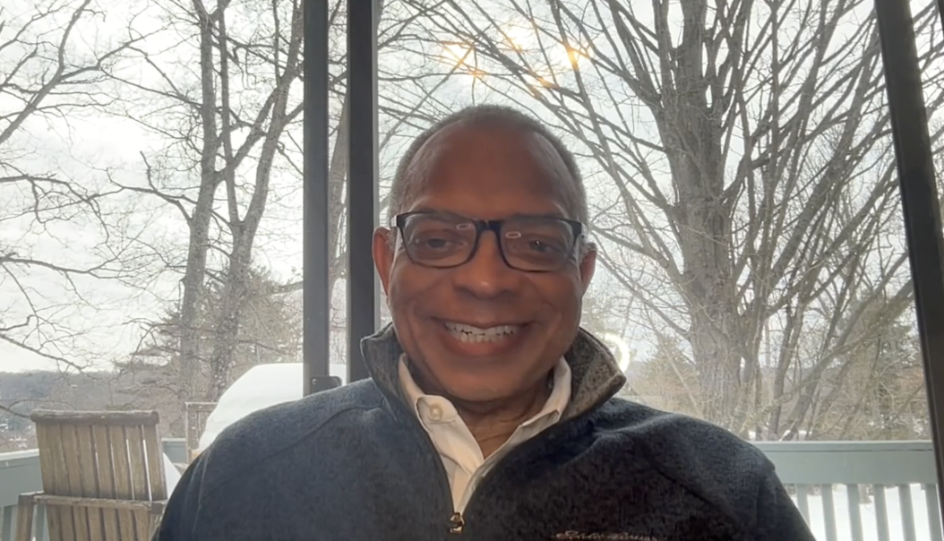 Man in glasses smiling indoors, wearing a dark jacket and white shirt. Background shows bare trees and a snowy landscape through a window.