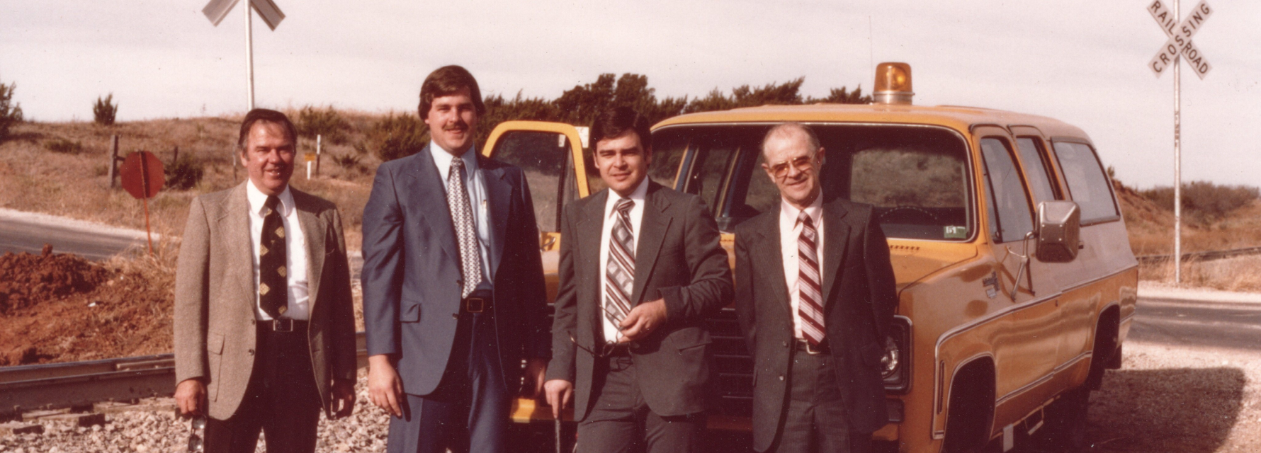 Four men in suits posed in front of a yellow service vehicle with rooftop beacon beside a rural railroad crossing.