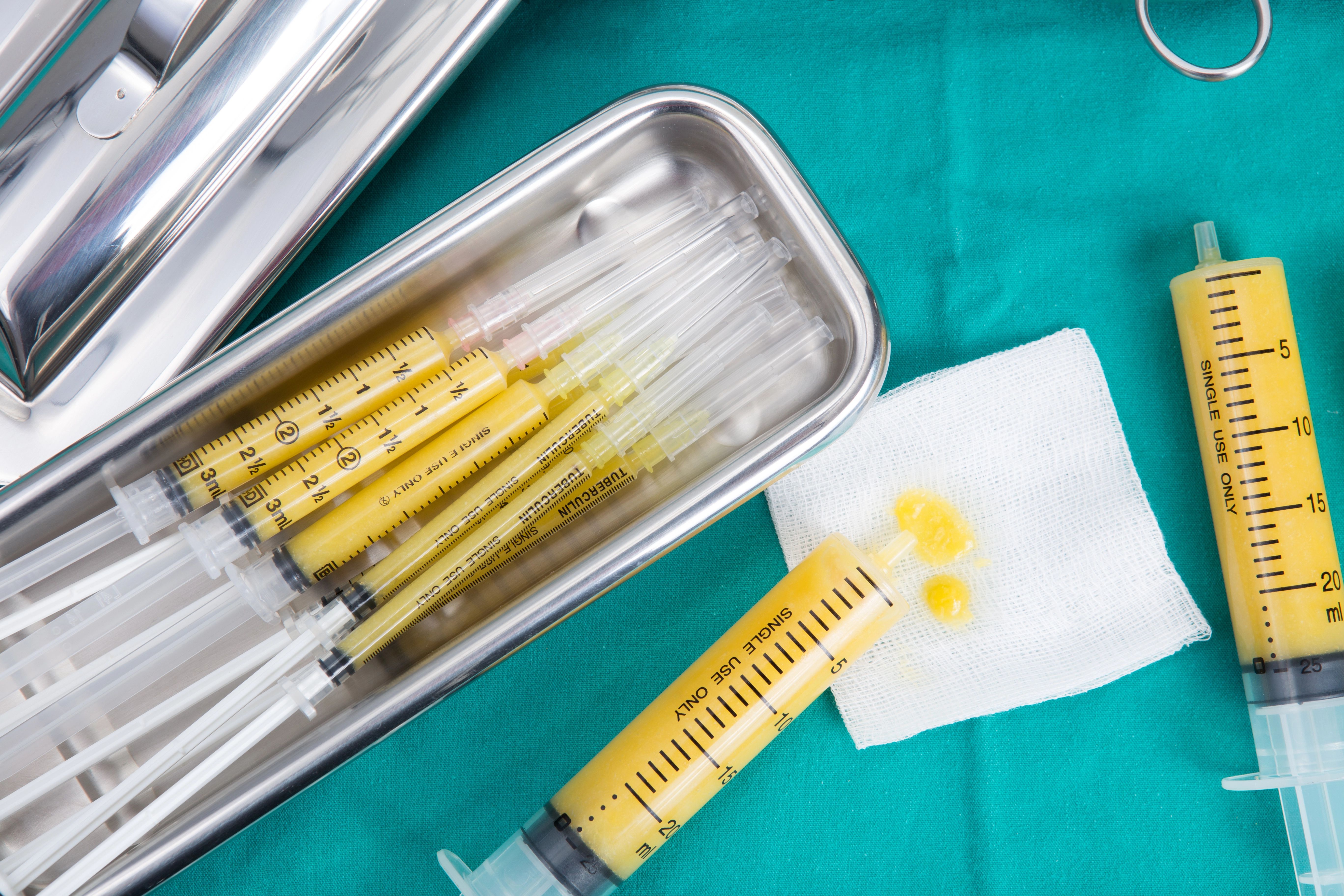 Syringes filled with yellow liquid in a metal tray, with one syringe and a gauze pad on a teal surface.