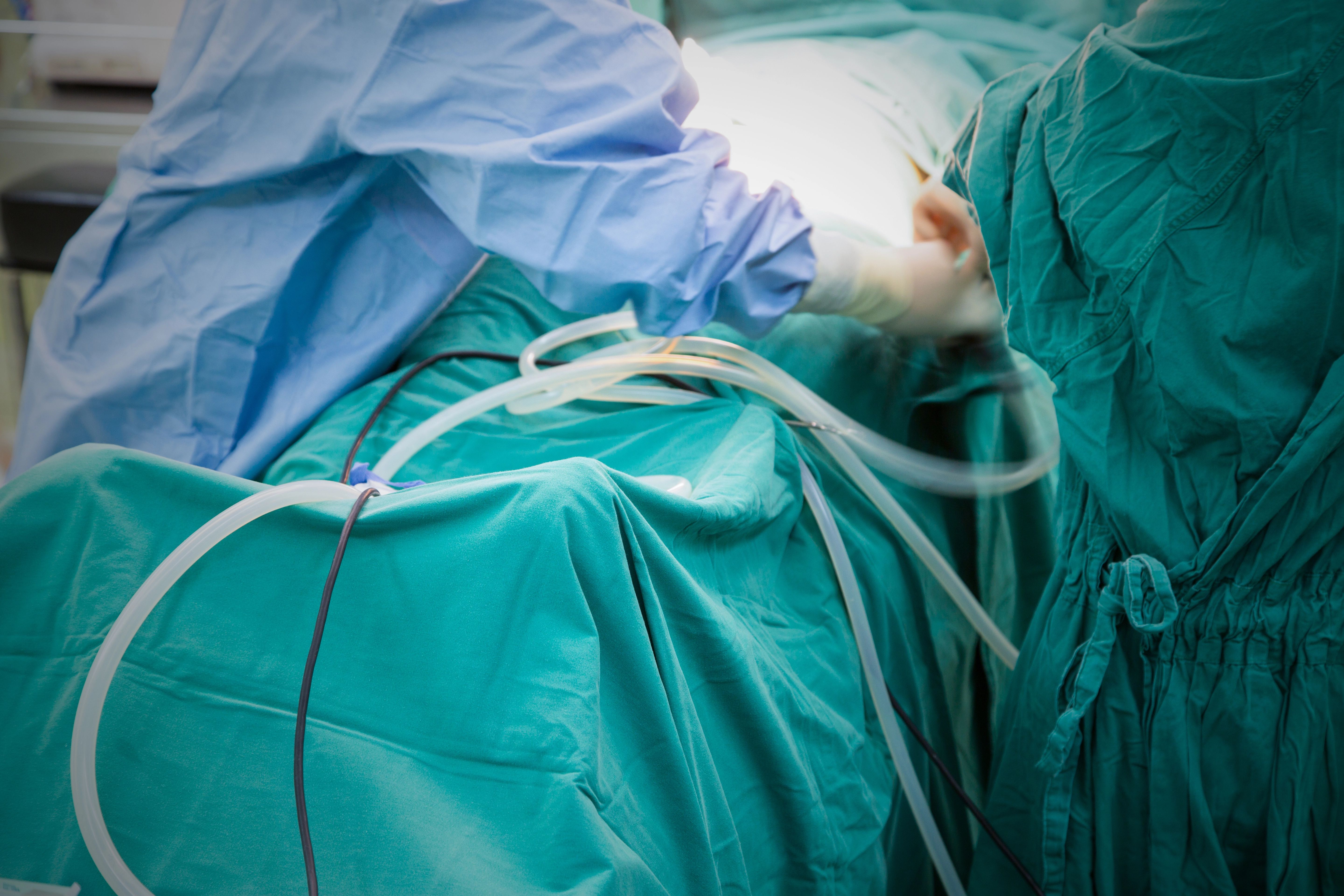 Surgeons in blue and green gowns perform an operation in a sterile, brightly lit operating room, with surgical instruments and tubes visible.