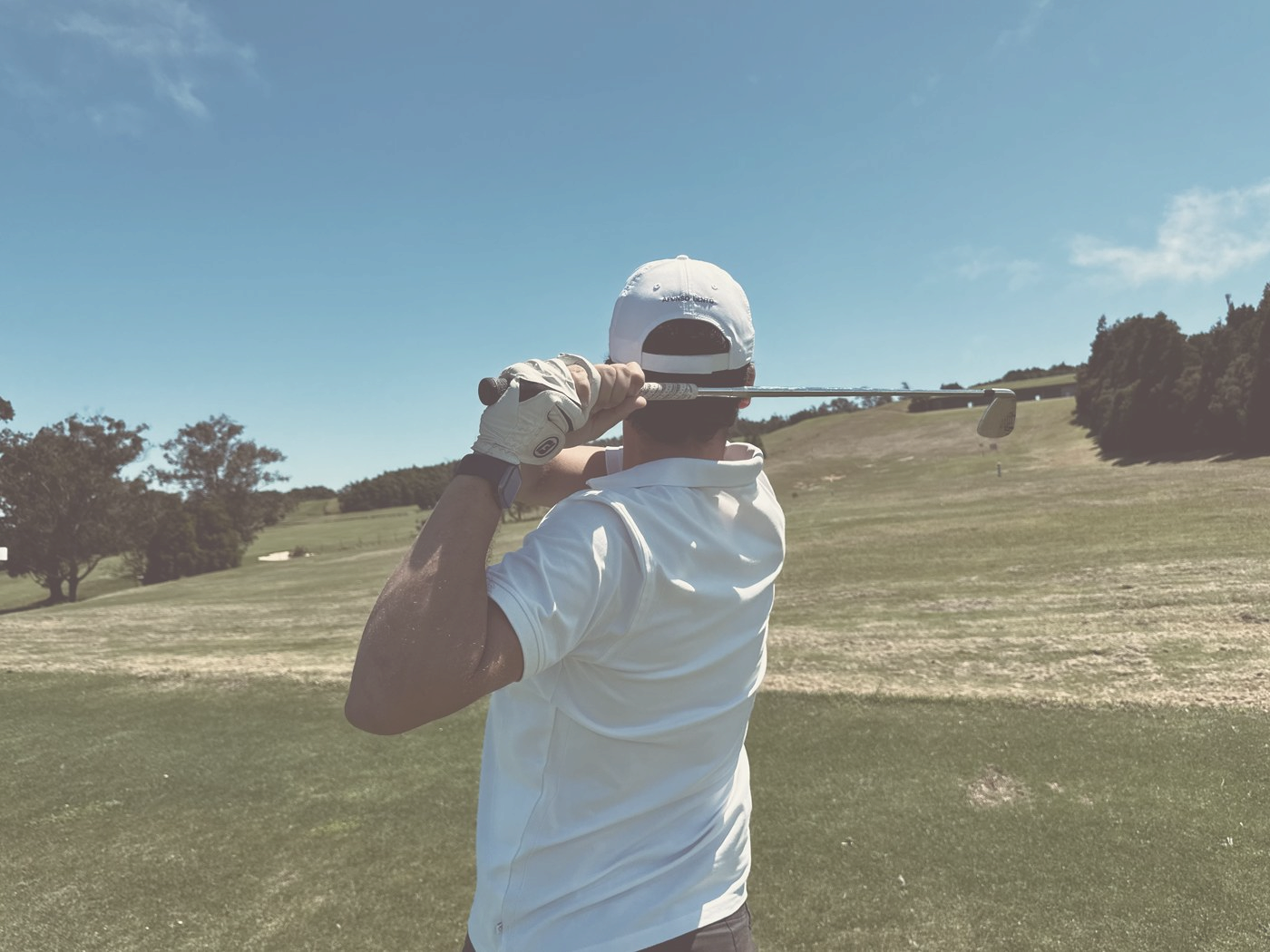 Golfer seen from behind finishing a swing on an open hilltop fairway, hazy warm light