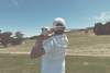Golfer seen from behind finishing a swing on an open hilltop fairway, hazy warm light
