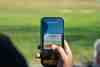 Golfer holding a phone showing the Hole19 app at a driving range