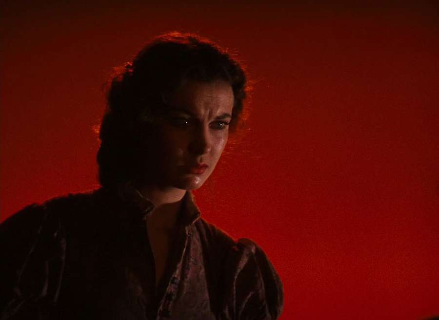 The image shows a person with dark hair and a serious expression, standing in front of a fiery red background.