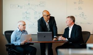 Man presenting to two others from a laptop with white board behind them