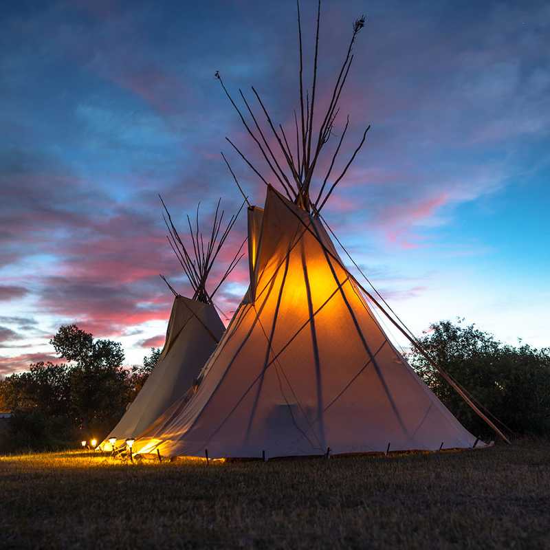 Canvas teepee illuminated at dusk with camp lights around it under a colorful twilight sky