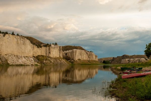 Calm river with a red dock and green shoreline beside limestone cliffs under a cloudy sky in Montana