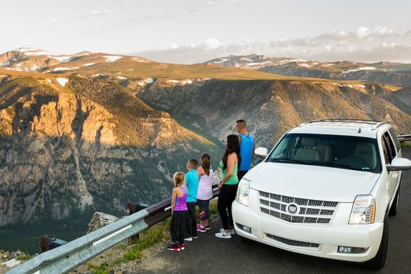 View of a family standing beside a mountain overlook road with a white car in the foreground and Montana hills and peaks in the background