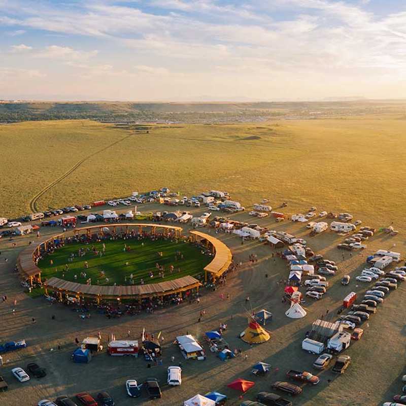 Aerial view of an outdoor car show parking lot in Montana with vehicles gathered around a central event area in open fields at sunset