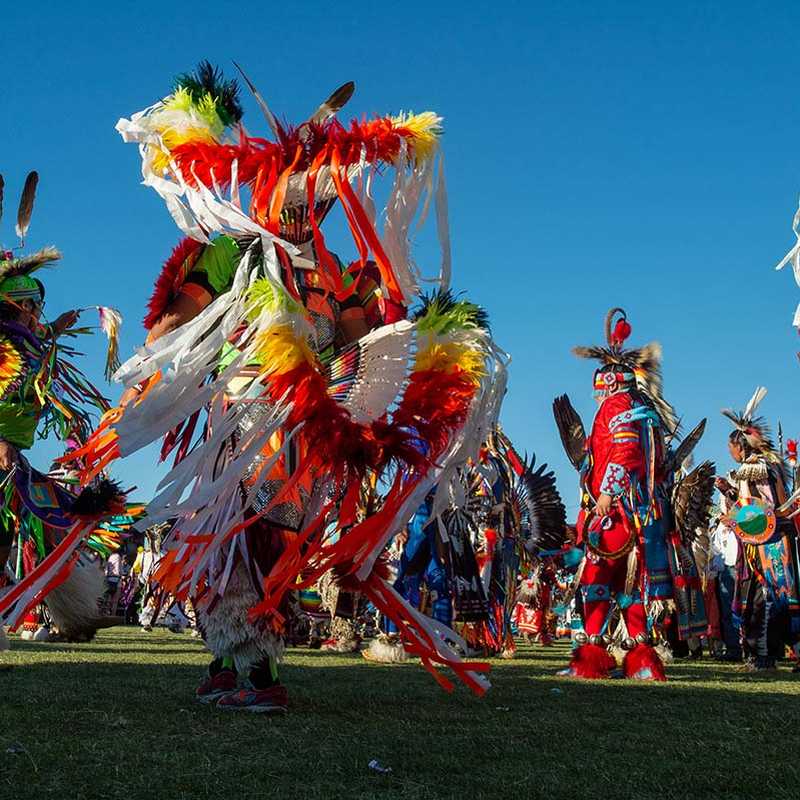 Native dancers in colorful regalia perform in a group outdoors under a clear blue sky