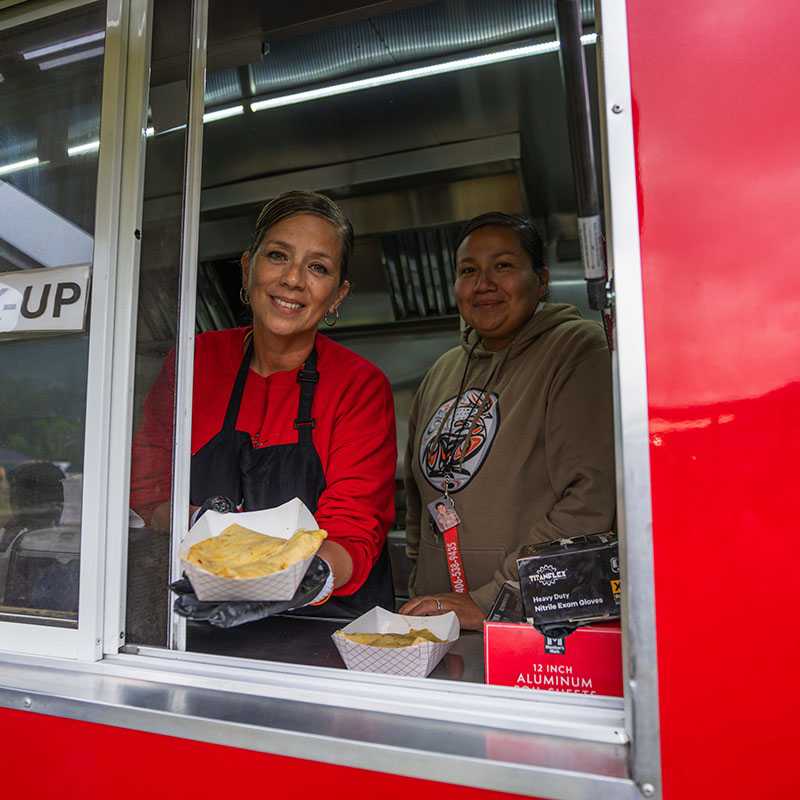 Two people smiling in a red food truck serving window holding prepared food in paper trays and bags