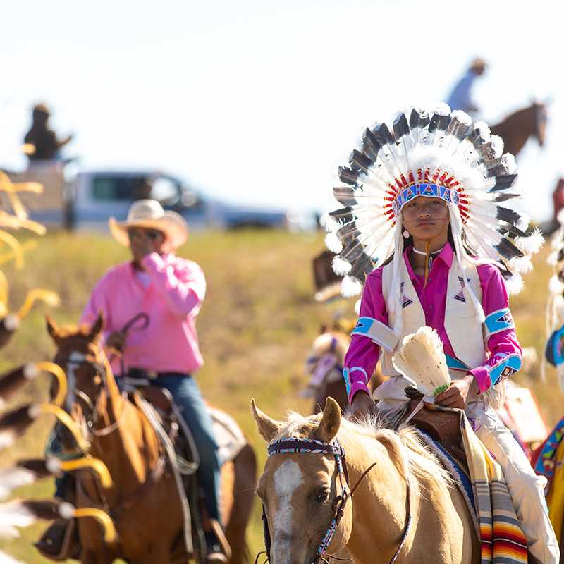 Native dancers in colorful regalia ride horseback at a powwow in Montana