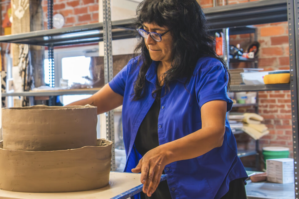 Person working at a workshop table, shaping materials at a workbench inside a brick-walled storage area