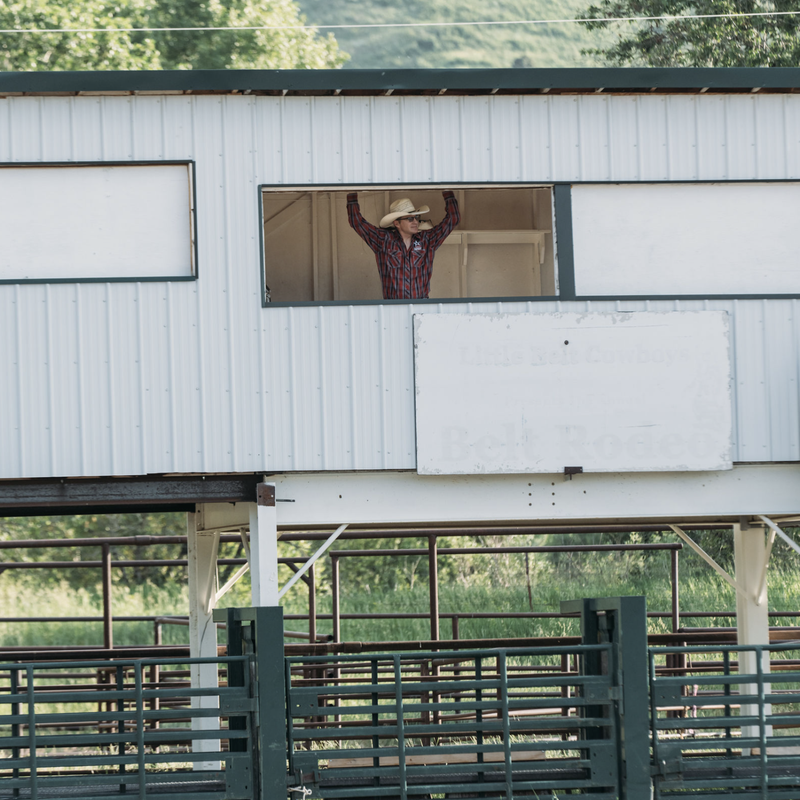 Person tipping a cowboy hat from inside an open doorway of a barn with green hills in the background