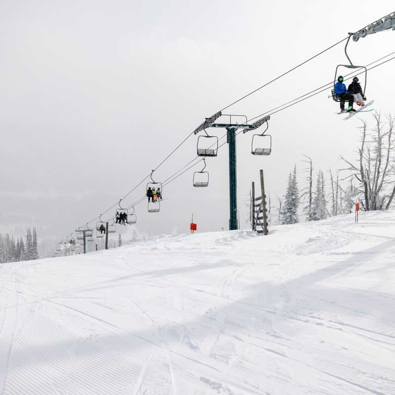 Skiers ride a chairlift above a snowy Montana slope with evergreen trees and lift towers in foggy weather