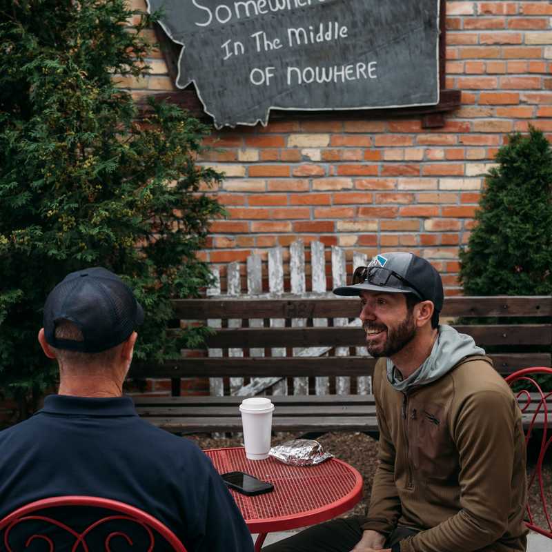 Two people sit at a red outdoor table beside a brick wall with a Montana-shaped sign reading “Somewhere in the middle of nowhere”