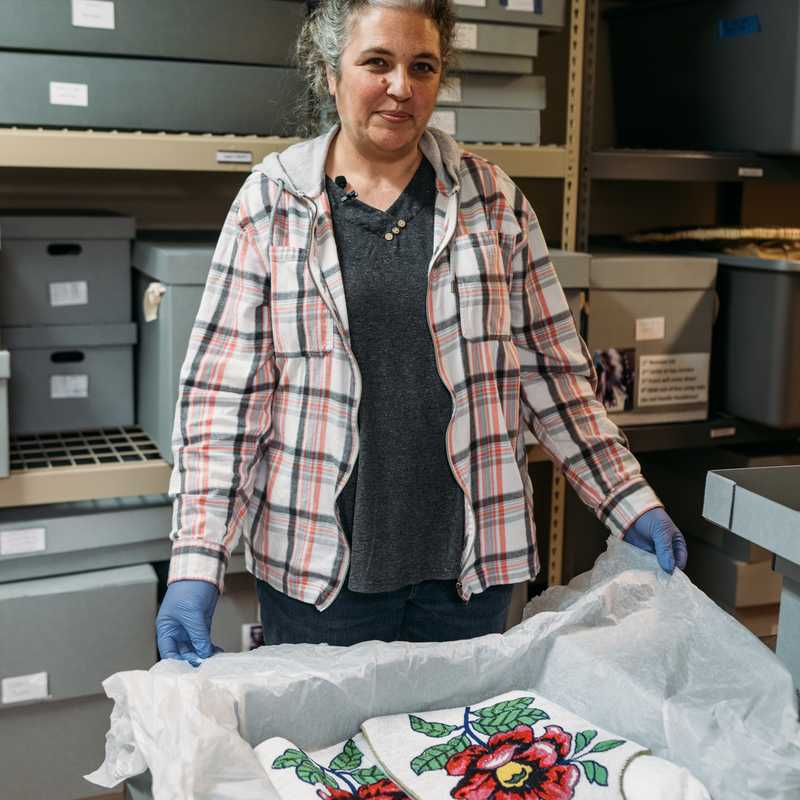 Person wearing blue gloves in a storage room with labeled shelving, standing behind a wrapped bundle with red and green floral fabric pattern