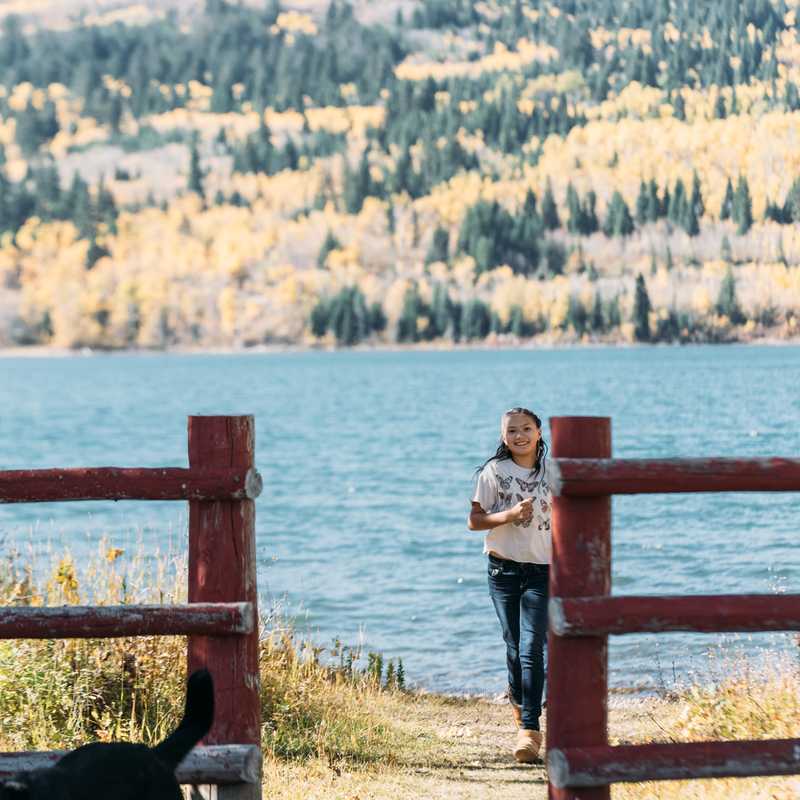 Woman walking along a lakeside path beside a fenced area, with a wide lake and forested hills in the background