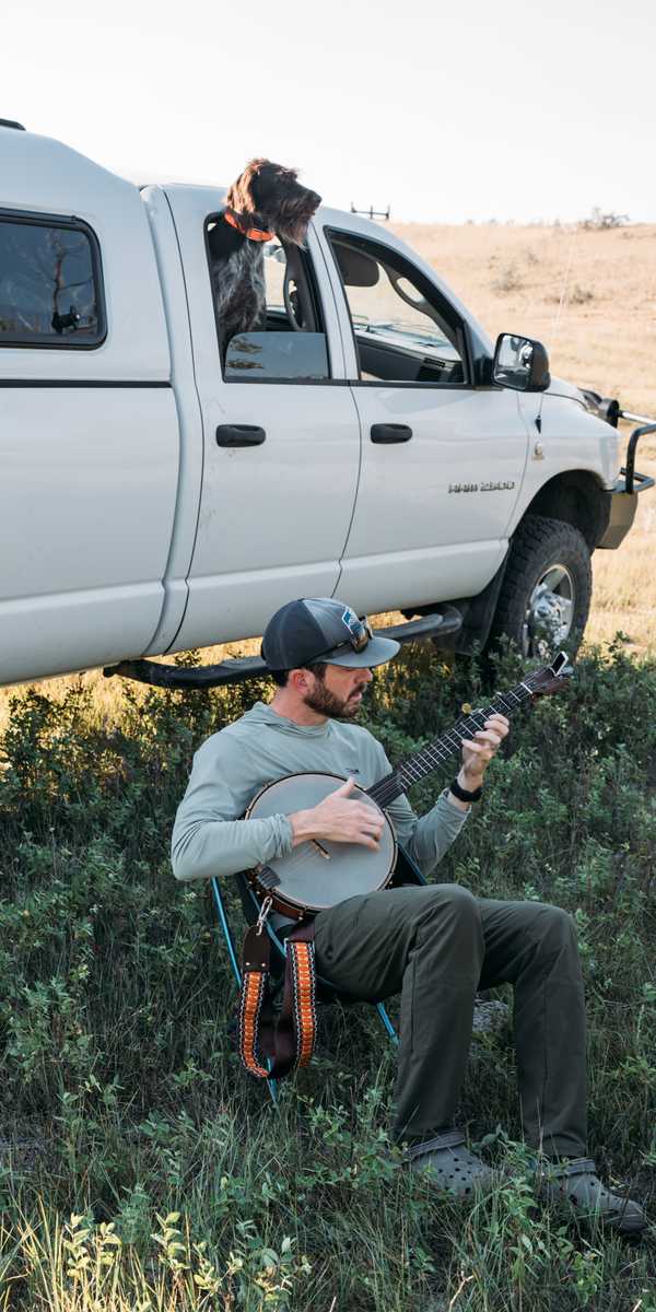 Musician playing a mandolin while relaxing outdoors near a white pickup truck in a rural Montana landscape