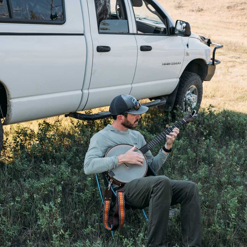 Musician playing a mandolin while relaxing outdoors near a white pickup truck in a rural Montana landscape