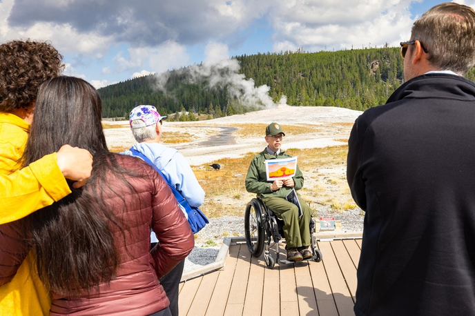 Tourists watch a performer on a boat-launch viewing platform while a river and forested hills sit in the background