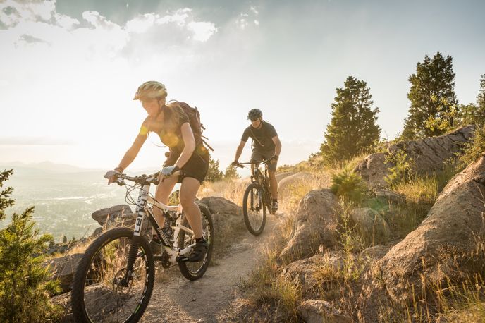 Two mountain bikers ride along a dirt trail with forested hills and a sweeping Montana valley view in the background