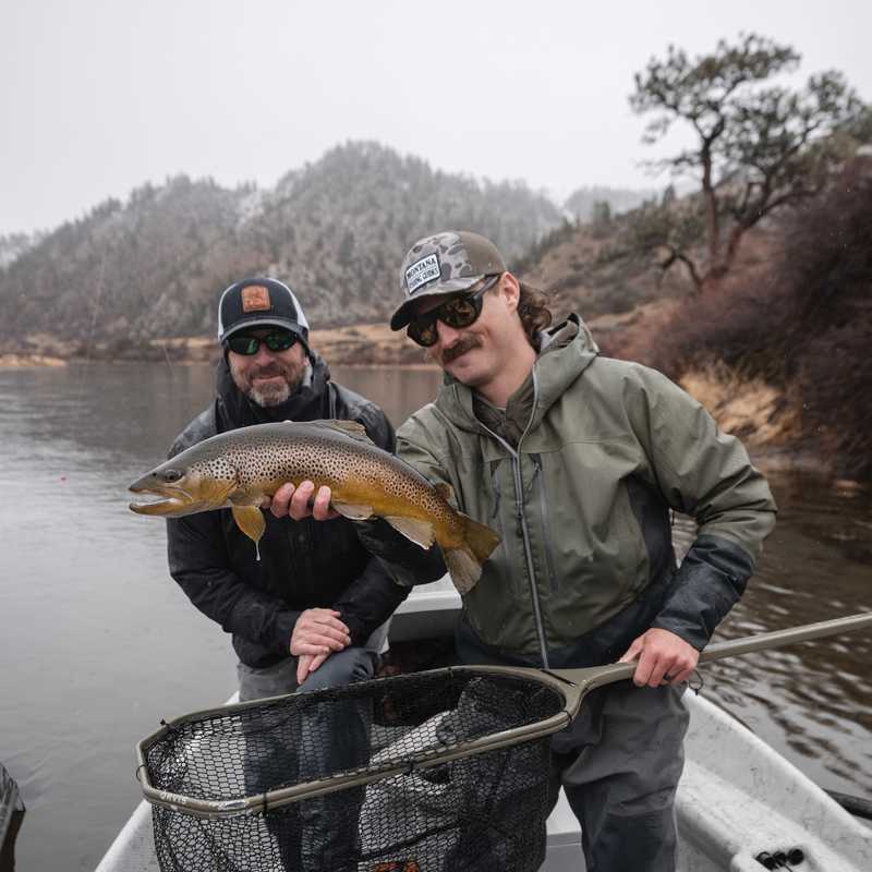 Two anglers hold a trout on a small fishing boat on a Montana river, with a landing net in the foreground