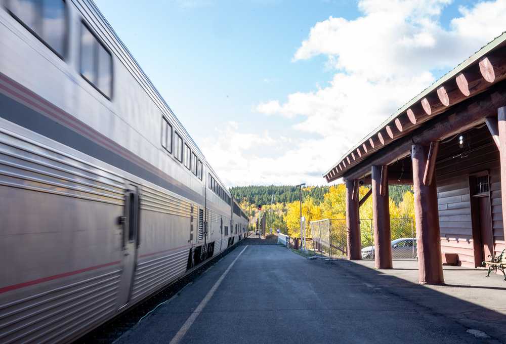 Passenger train passing through train station in Montana with fall foliage in background.