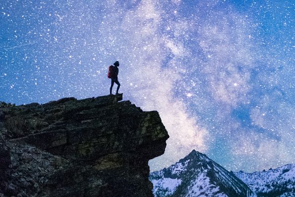 Person standing on a rocky overlook at night with the Milky Way over snowy Montana mountains