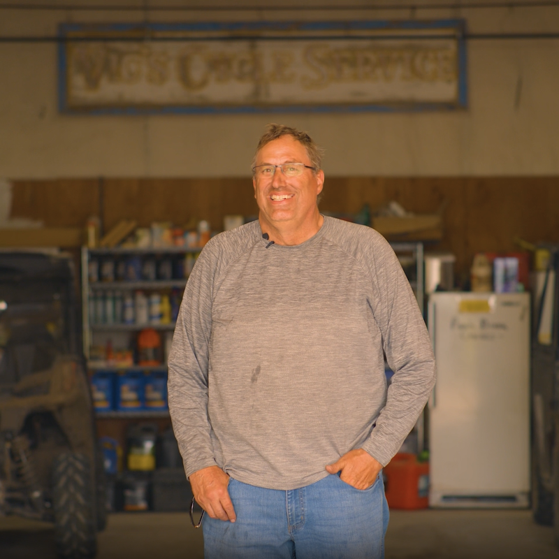 Man standing indoors at a workshop or service bay with tools, equipment, and vehicles in the background