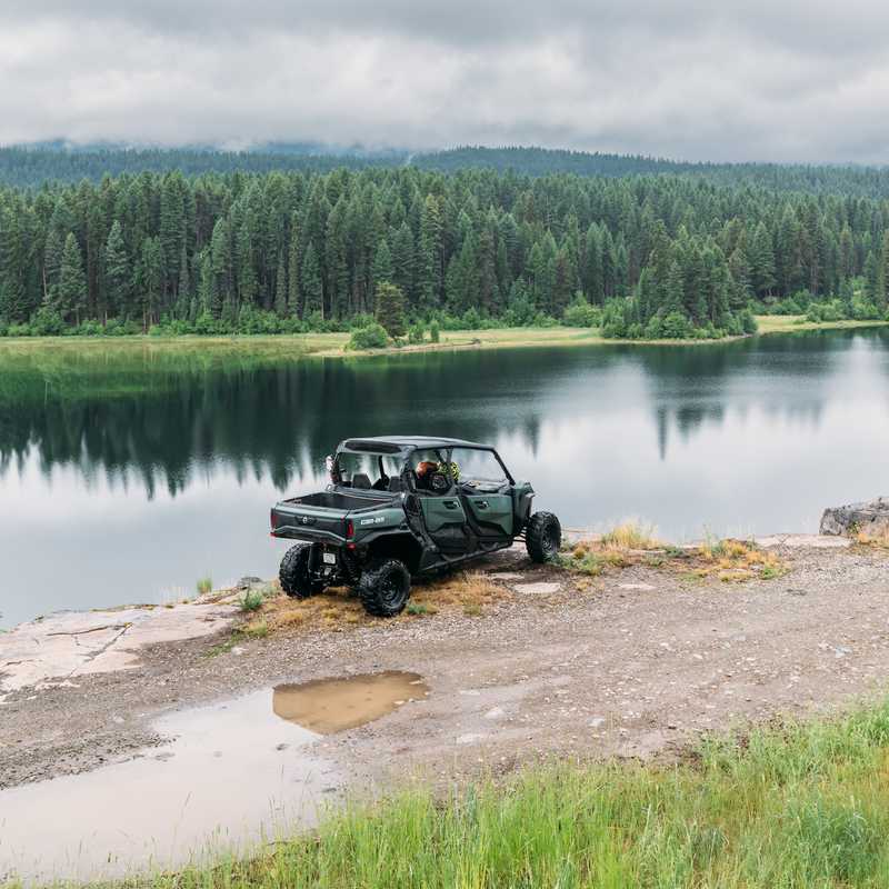 Off-road UTV parked by a calm lake with forested hills under an overcast sky in Montana