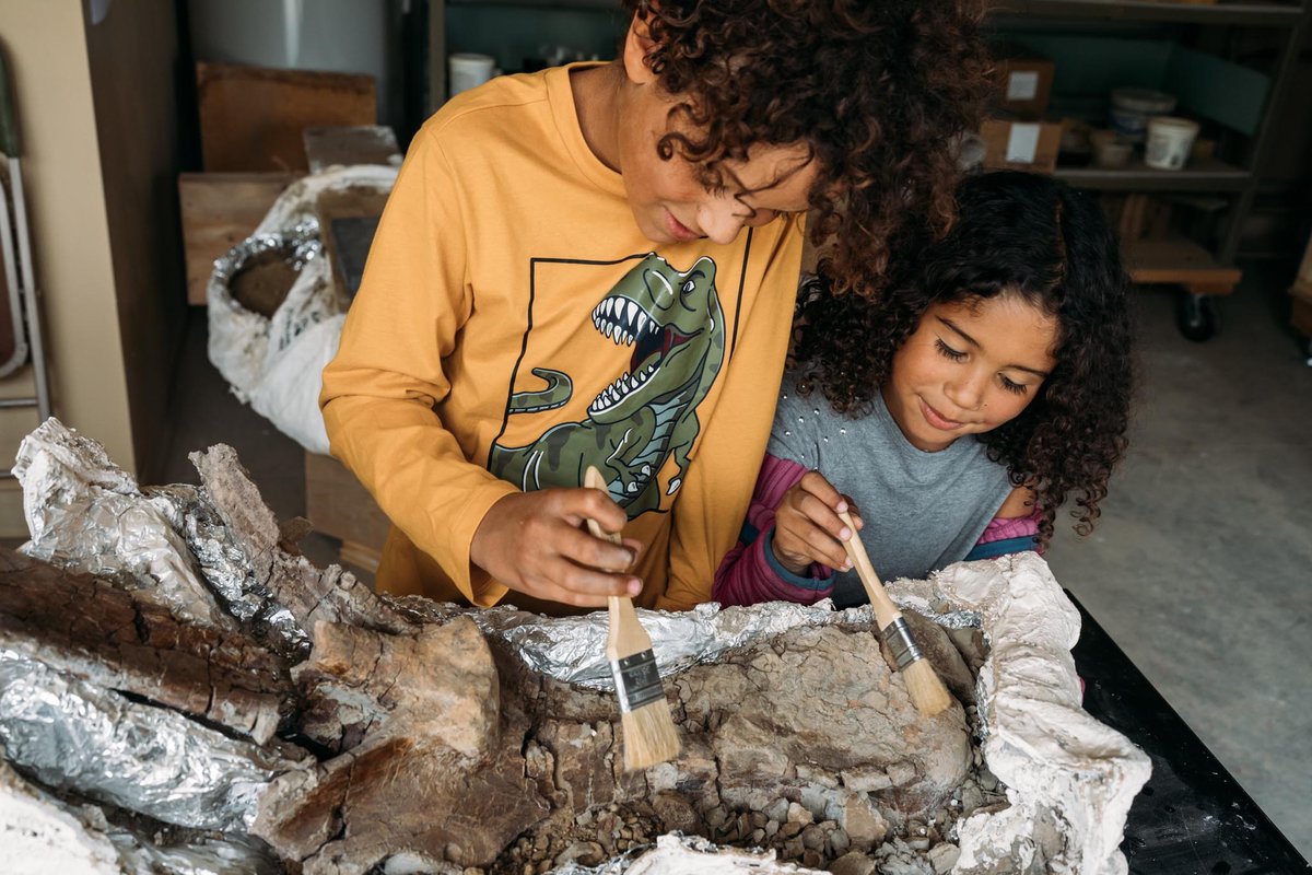 Two people carve and decorate woodcut figures at a workbench, with tools and protective materials nearby